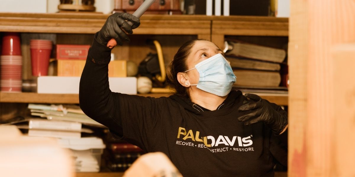 A Paul Davis Restoration specialist wearing a surgical face mask and black protective gloves using a tool to inspect shelves filled with household items.